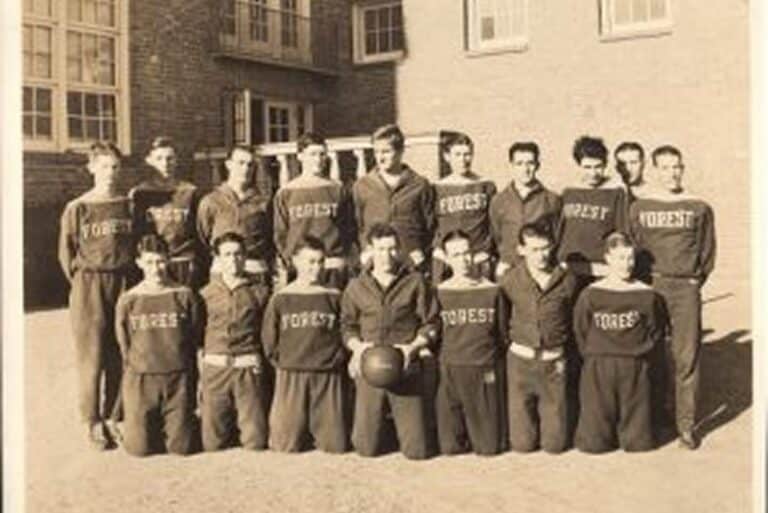 Vintage photo of a boys’ basketball team in matching “Forest” uniforms posing outside a brick building.