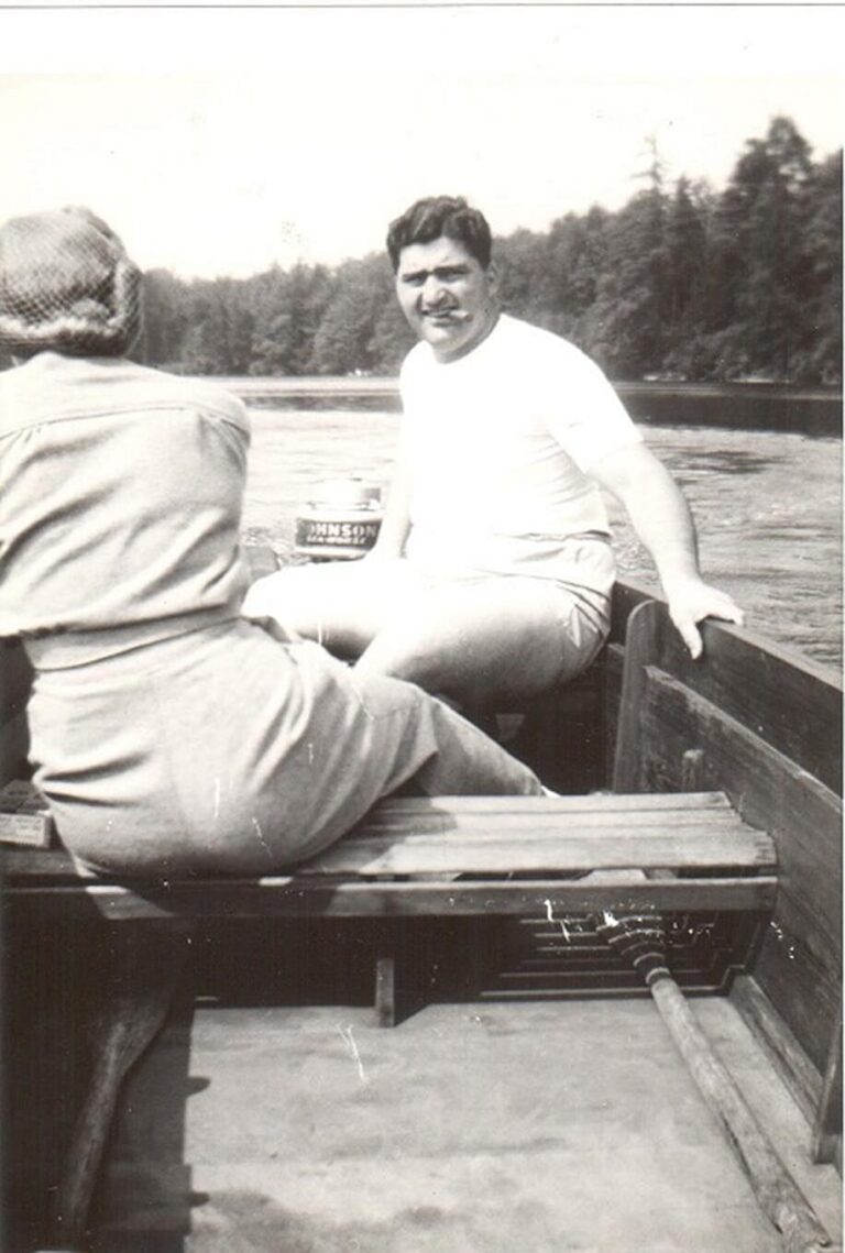 Two people sit in a small rowboat on a river, surrounded by trees, on a sunny day.