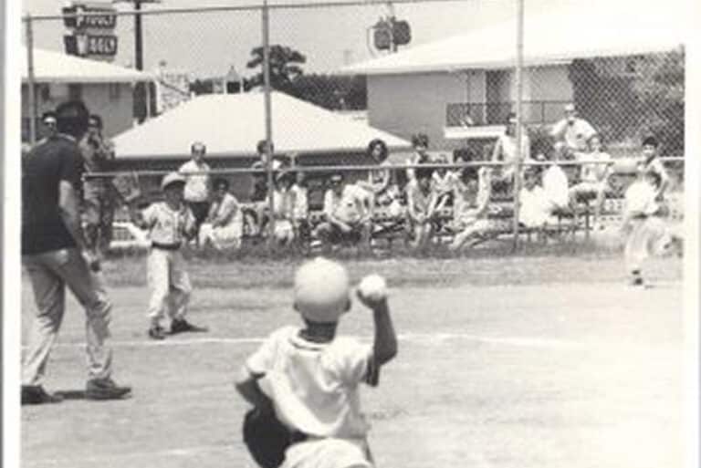 A child throws a baseball to a batter while people watch from bleachers on a sunny day at a ballpark.