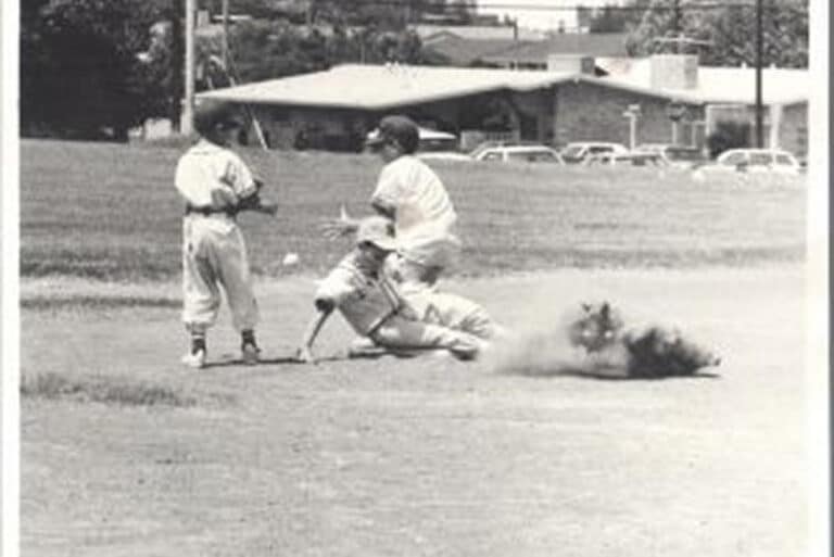 Three boys play baseball; one slides into base, raising dust, while another tries to tag him, and a third looks on.
