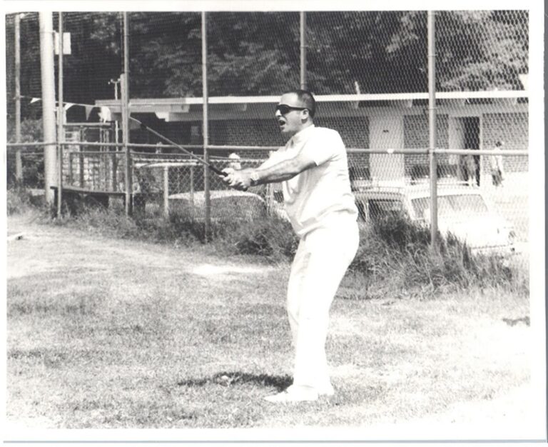 Man in sunglasses and white clothes holds a baseball bat, preparing to swing on a grass field near a chain-link fence.