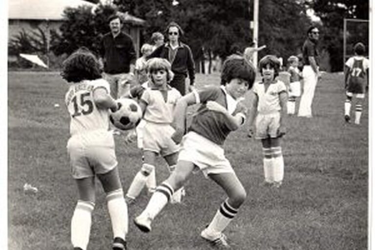 Children play soccer on a grassy field while adults watch in the background. Players wear numbered uniforms and athletic socks.