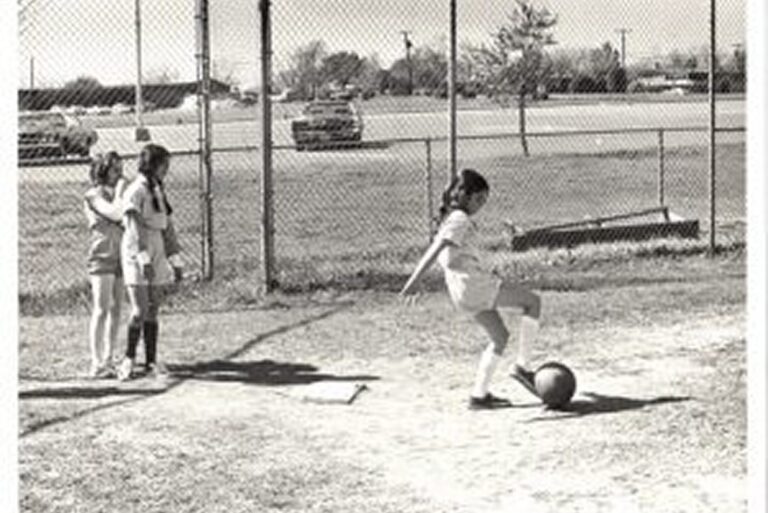 A girl about to kick a soccer ball while two others watch near a chain-link fence on a grassy field.