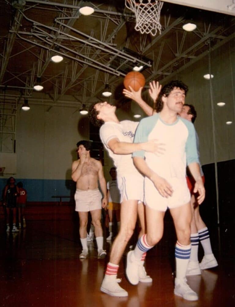 Four men in vintage basketball uniforms play under a hoop in a gym, one attempting a close shot.