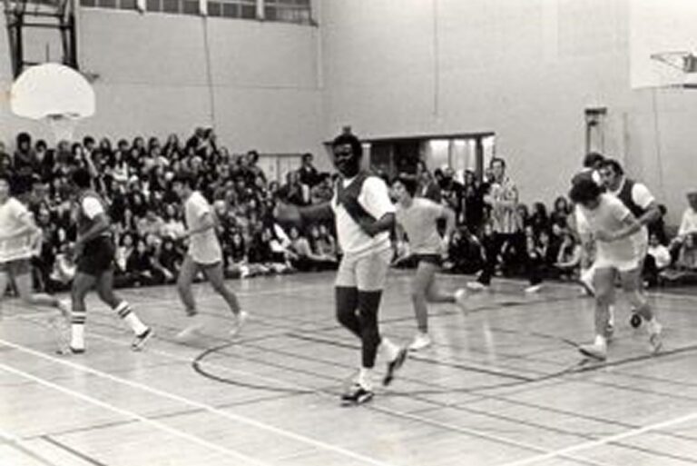 A group of people play basketball in a gym while a crowd of spectators watches from the bleachers.