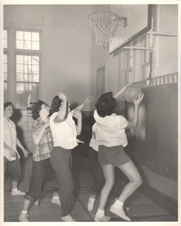 Four women play basketball indoors; one prepares to shoot while others defend under the hoop.