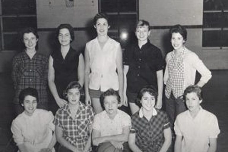 Ten young women in casual shirts pose together indoors in a black-and-white group photo, some standing, some seated.
