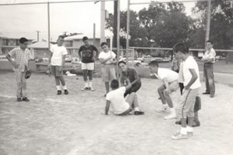 A group of boys play baseball on a dirt field, with one boy sitting on the ground near home plate.