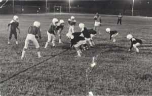 Children wearing football gear line up on a field at night, preparing for a play under bright stadium lights.