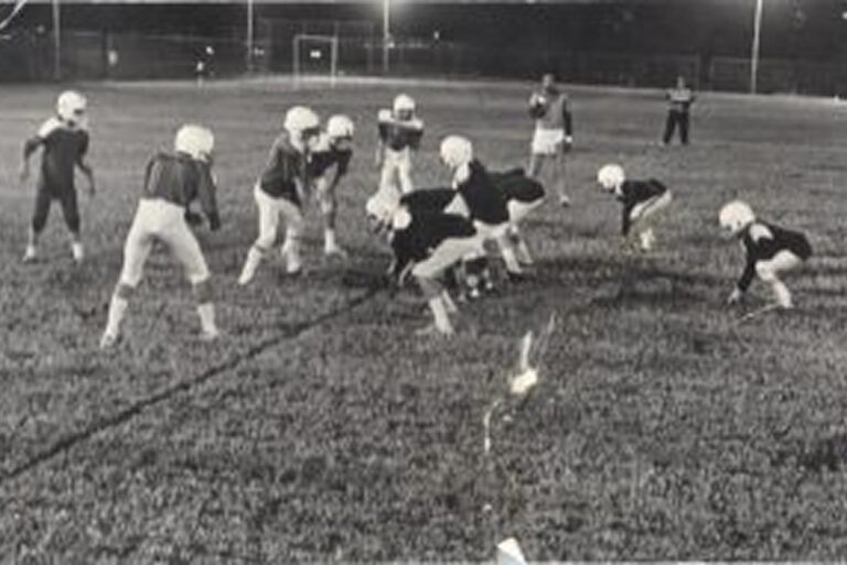 Youth football players in helmets and uniforms line up on a field at night, preparing to start a play.