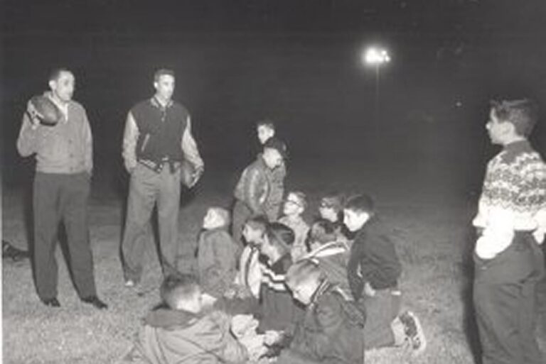 Two adults stand with a football while a group of boys sit and listen on a grassy field at night.