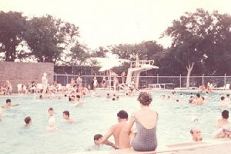 Crowded public swimming pool with children and adults; a diving board and trees are visible in the background.