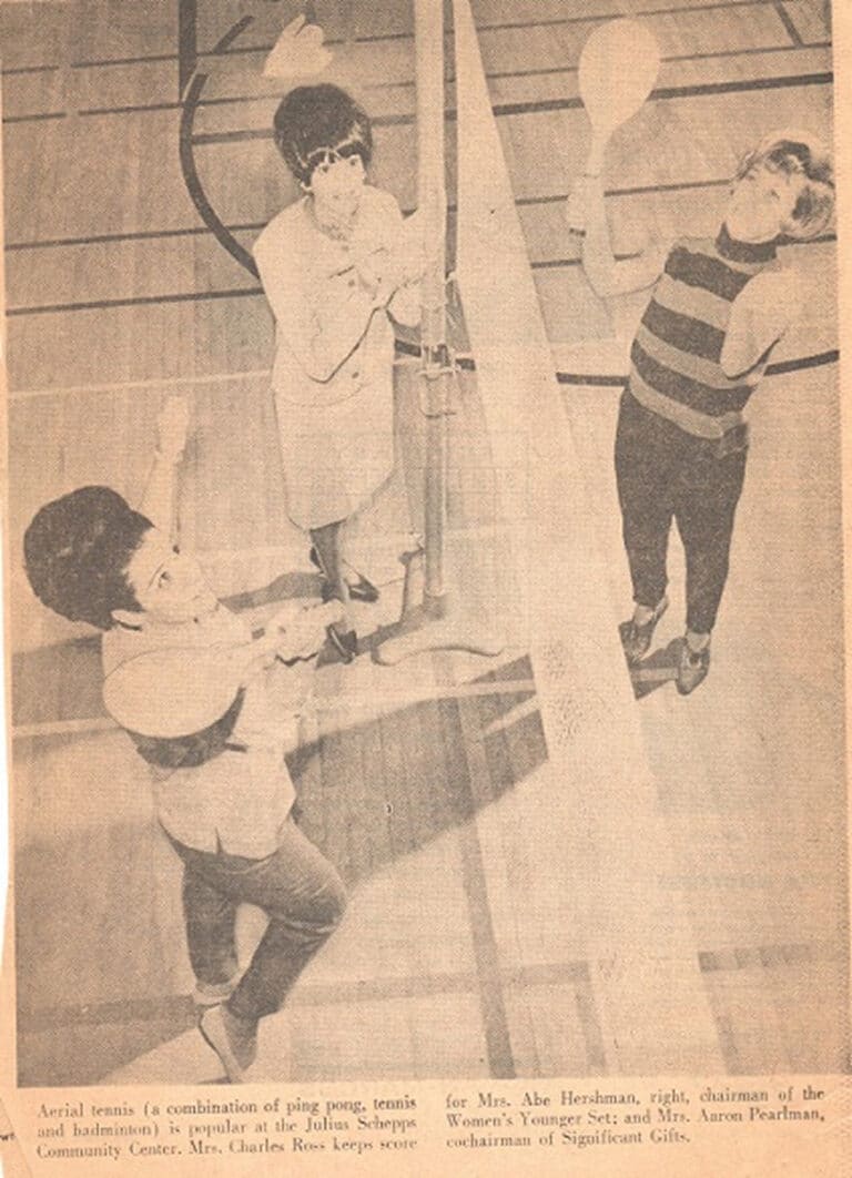 Three women play a paddle game over a net in a gym, looking up at a flying ball or birdie.