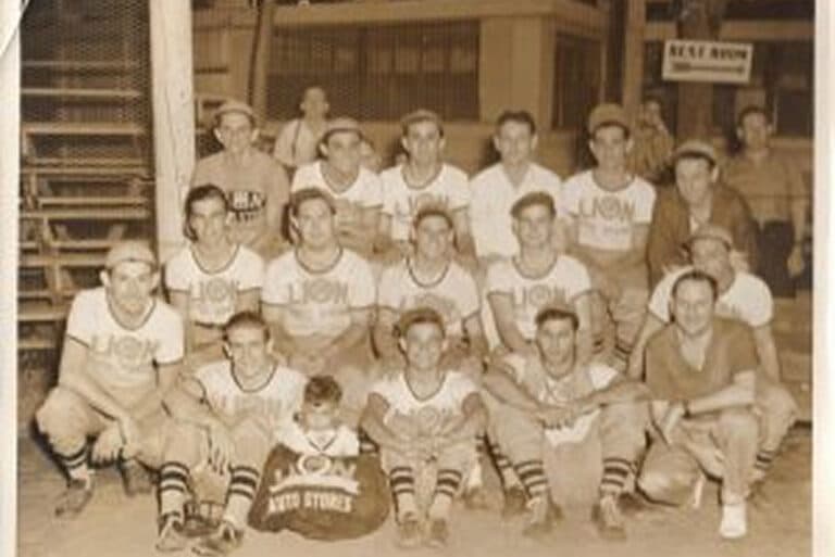 Vintage photo of a baseball team in uniforms posing together; one child sits in front holding a team bag.