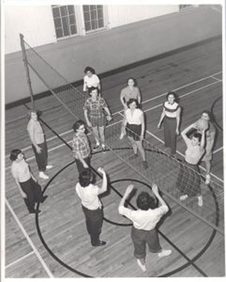 Black and white photo of people standing on a gym floor around a volleyball net, looking up.