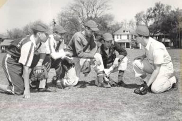Five baseball players kneel on a field in uniforms, appearing to discuss a play during daytime.