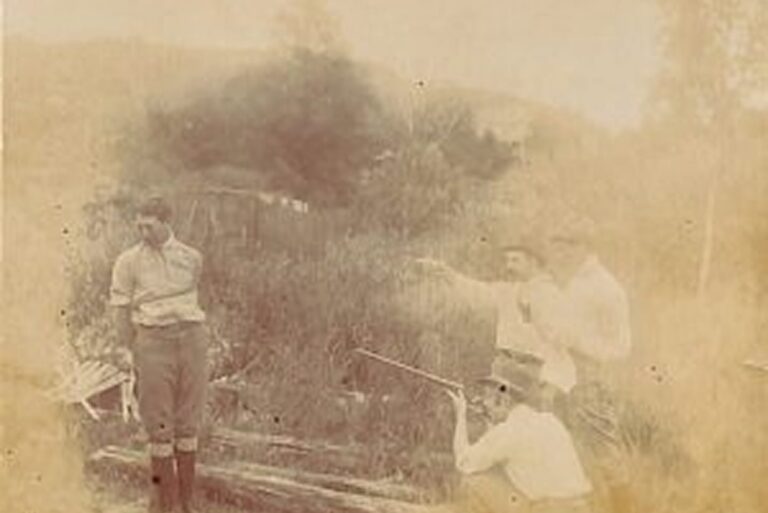Four men outdoors; one stands against a post while three others aim or point rifles toward him in a grassy, rural area.