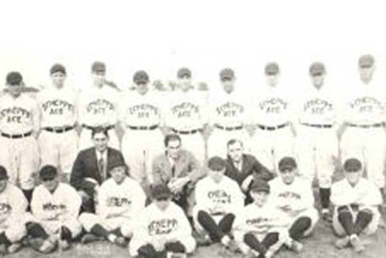 Vintage black and white photo of a baseball team posing in uniforms, with some standing and others sitting on the ground.
