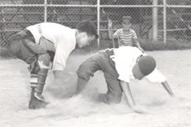 Two boys play baseball on a dusty field while a young child watches from behind a fence in the background.