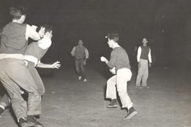 Four young men play a game outside at night, running and reaching toward each other on a dimly lit field.