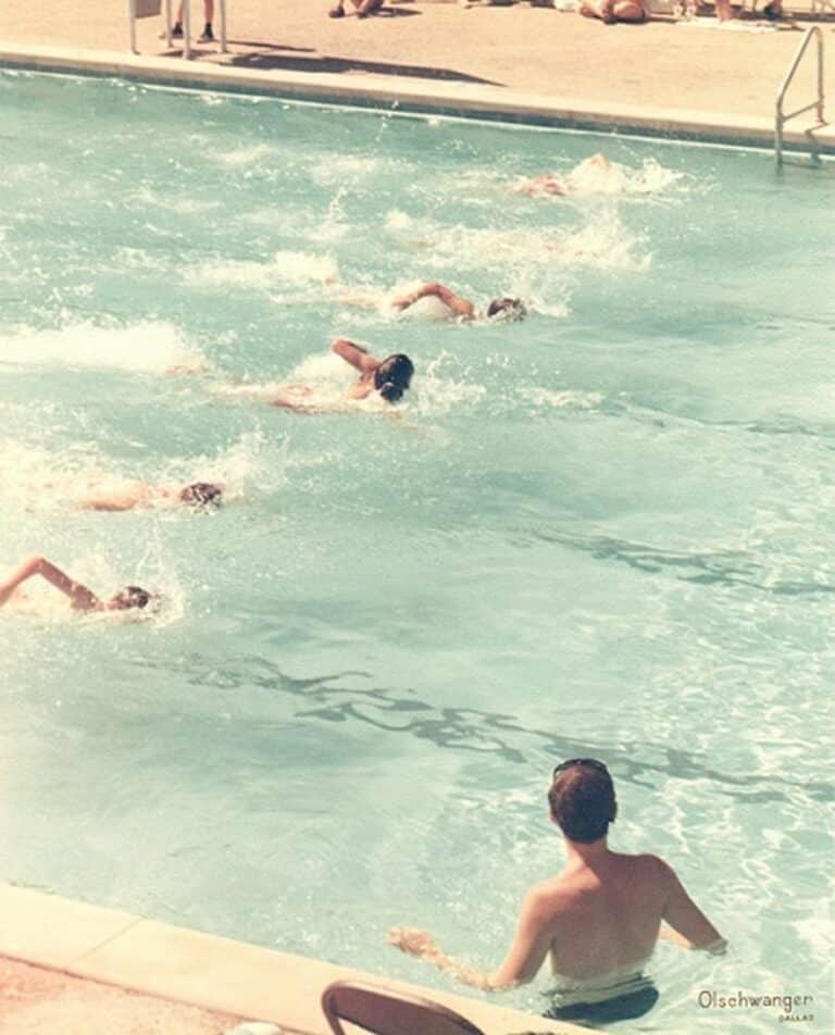 Several swimmers race in a pool while a person at the edge watches. Sunlight reflects off the water’s surface.