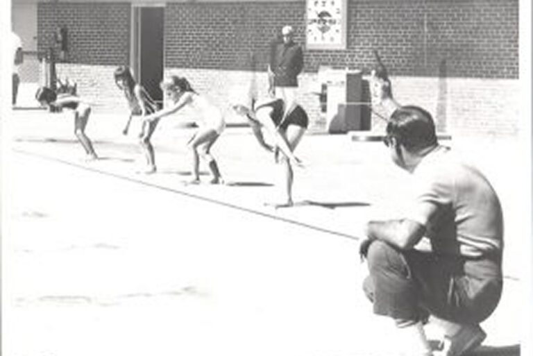 Five children in swimsuits prepare to dive into a pool as two adults watch, one crouching beside the pool.