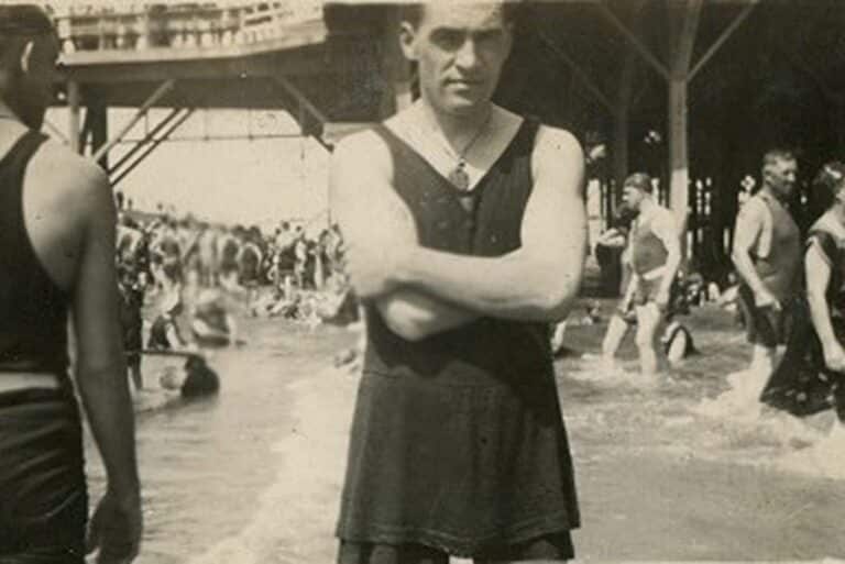 Man in vintage swimsuit stands with arms crossed on a crowded beach; people swim in the background under a pier.