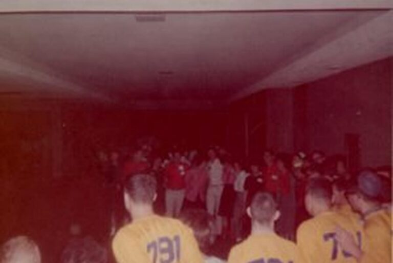 A group of people in yellow and red shirts gather in a dimly lit room, some facing away from the camera.