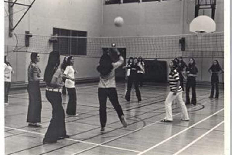 A group of people play volleyball indoors in a gym, with one person jumping to hit the ball over the net.