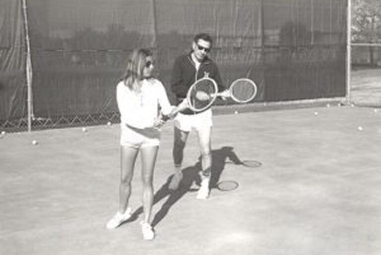 A man coaches a woman on a tennis court, both holding rackets, with tennis balls scattered on the ground.