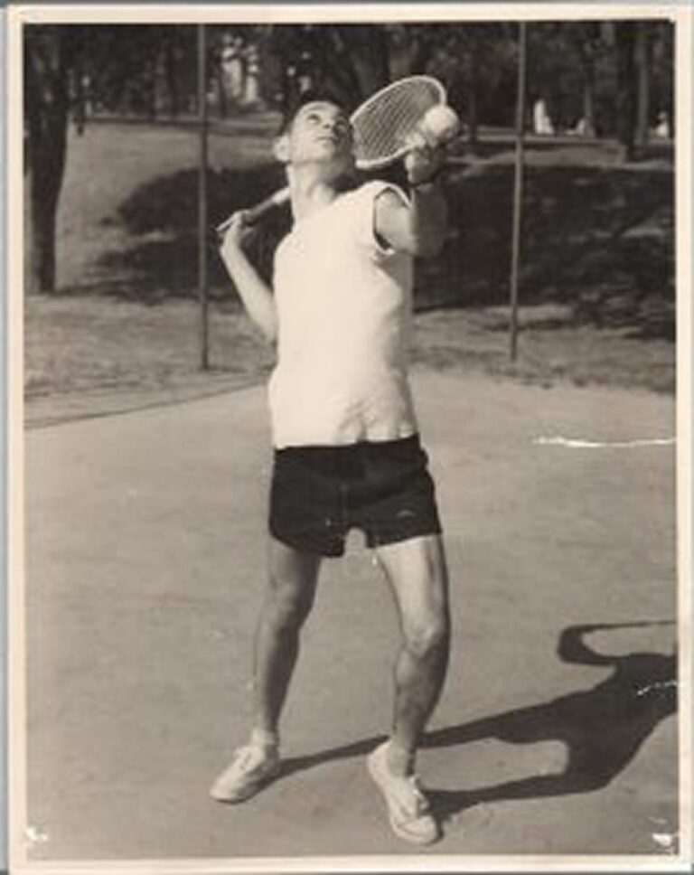 Young man in shorts and t-shirt prepares to hit a tennis ball with a racket on an outdoor court.