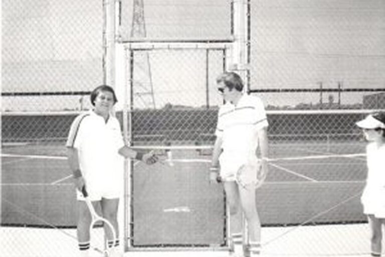 Three people in tennis attire stand by a chain-link fence on a tennis court, two holding rackets, one looking on.