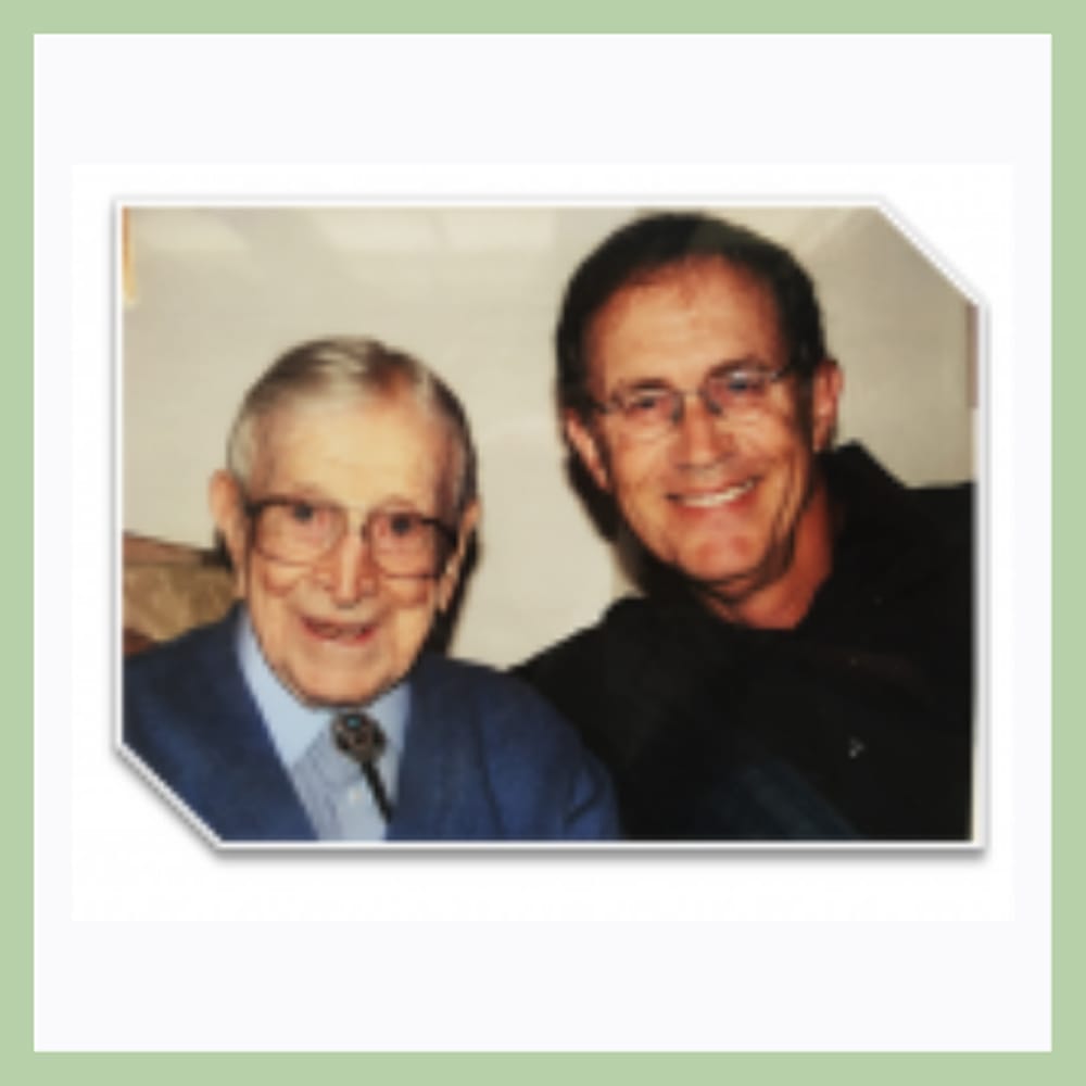 Two smiling men, one elderly in glasses and a blue sweater, one middle-aged, pose together for a photo.