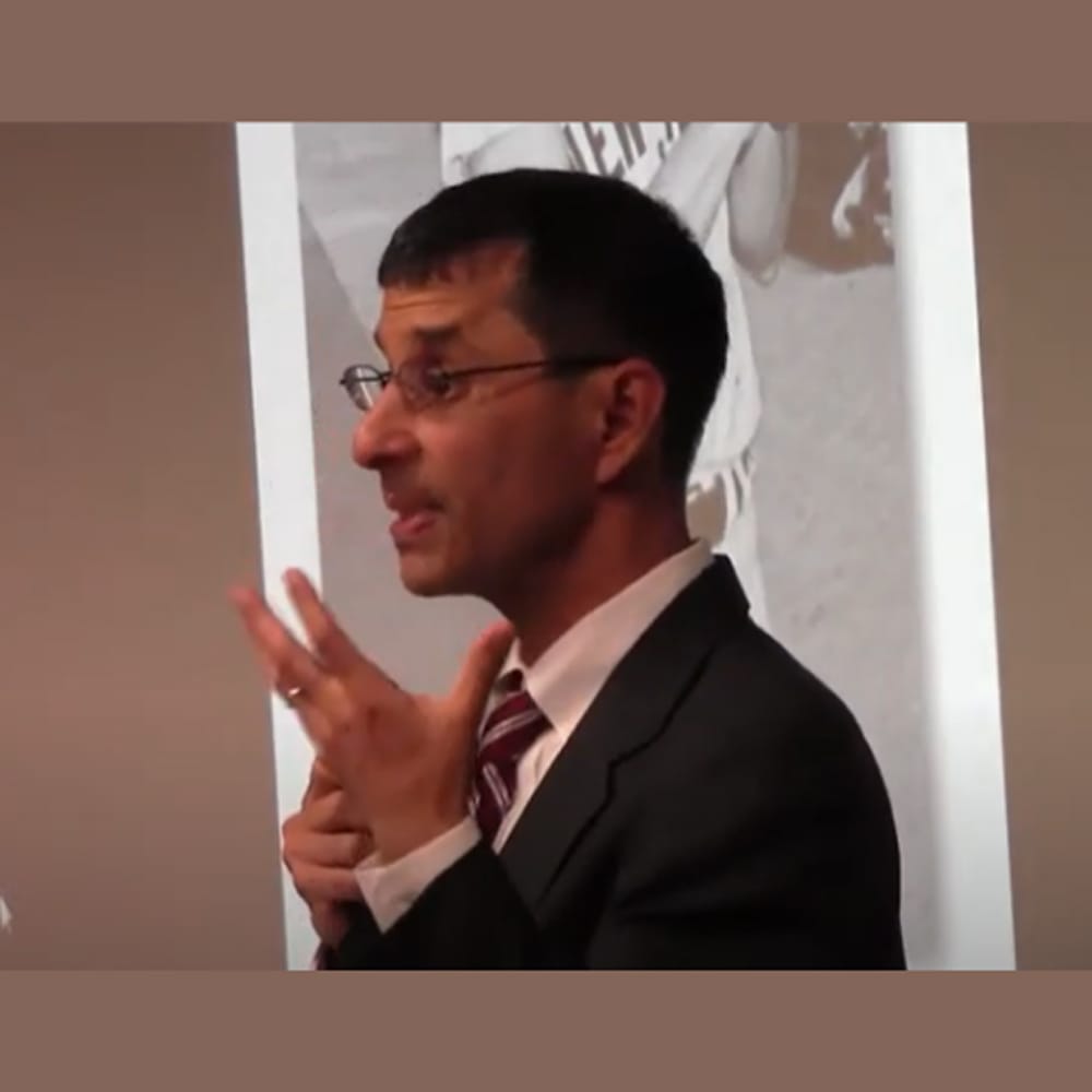 Man in a suit and glasses gestures while speaking, with a black-and-white photo projected behind him.