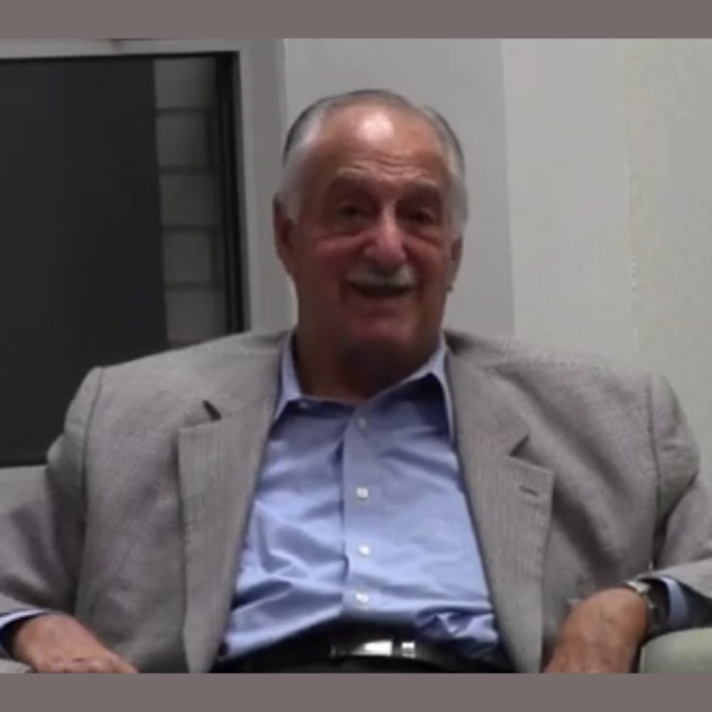 Older man in a gray blazer and blue shirt sitting indoors, smiling at the camera.