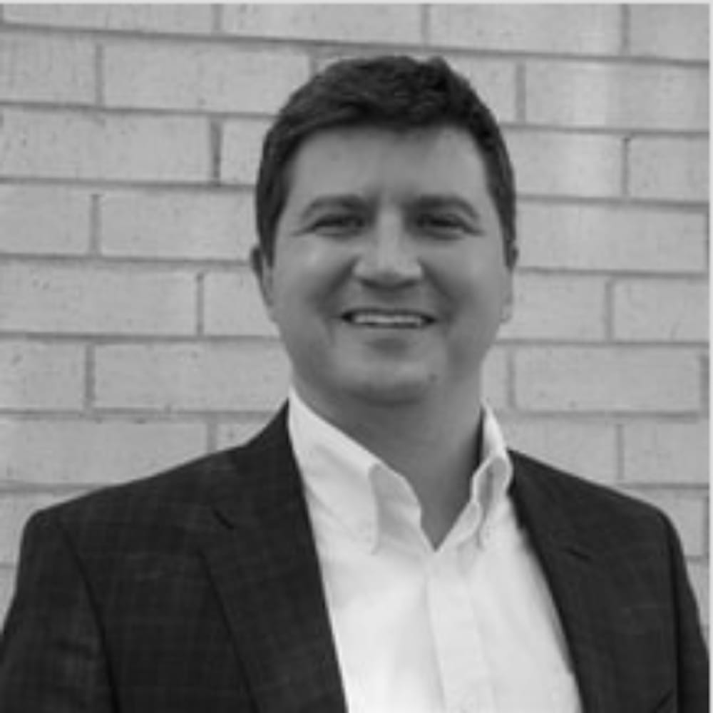 Man in a suit jacket and white shirt smiling in front of a light-colored brick wall, black and white photo.
