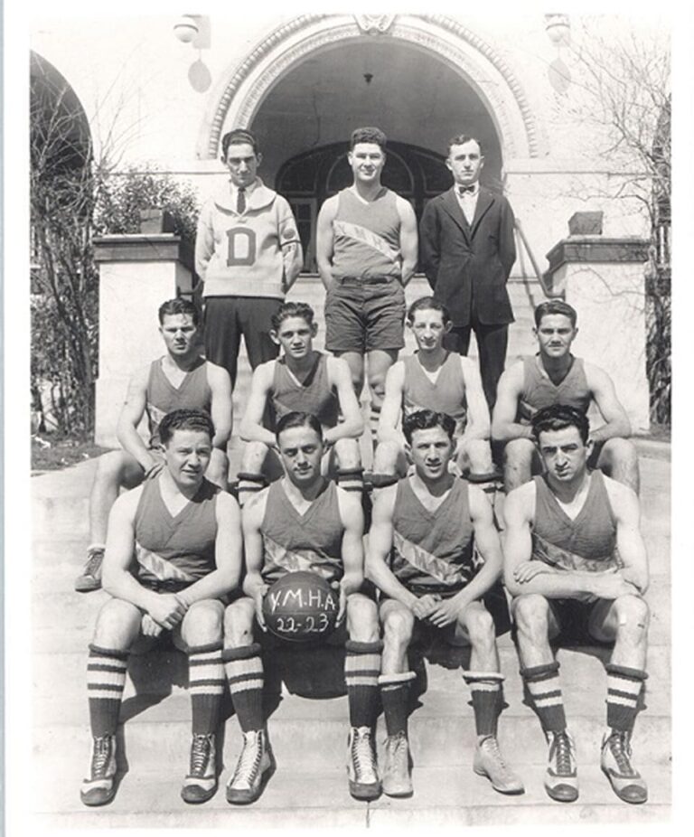 Vintage basketball team poses on steps; ten players in uniforms, two men in suits, holding “Y.M.H.A. 22-23” basketball.