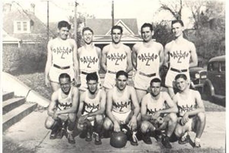 Black and white photo of a vintage basketball team posing outdoors, all wearing matching uniforms labeled “Dallas.”.