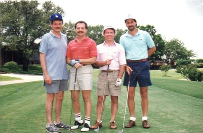 Four men in golf attire stand together on a golf course, smiling at the camera on a sunny day.