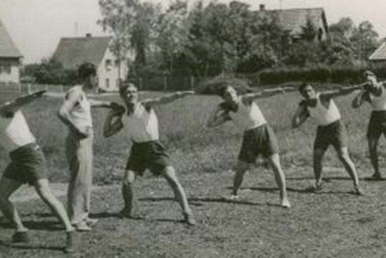 Six people in shorts and tank tops practice stretching exercises outdoors, with a coach standing nearby.