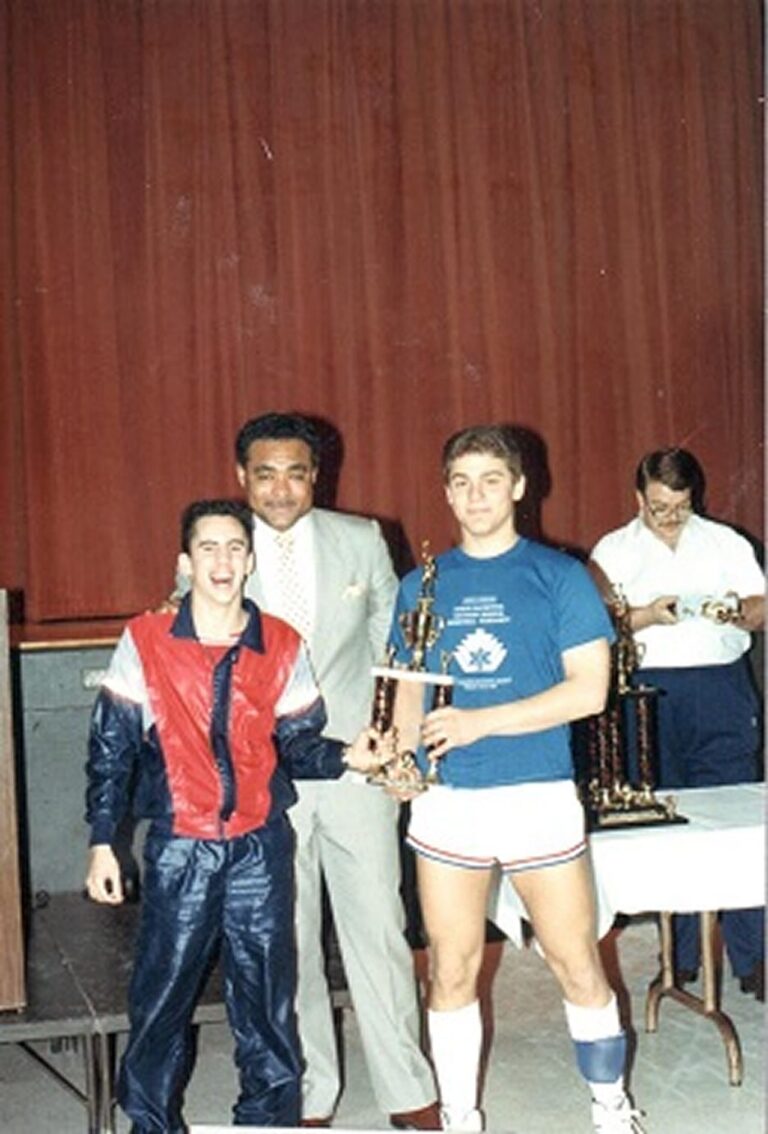 Three men stand together, two holding a trophy, with a third man behind a table with more trophies in the background.