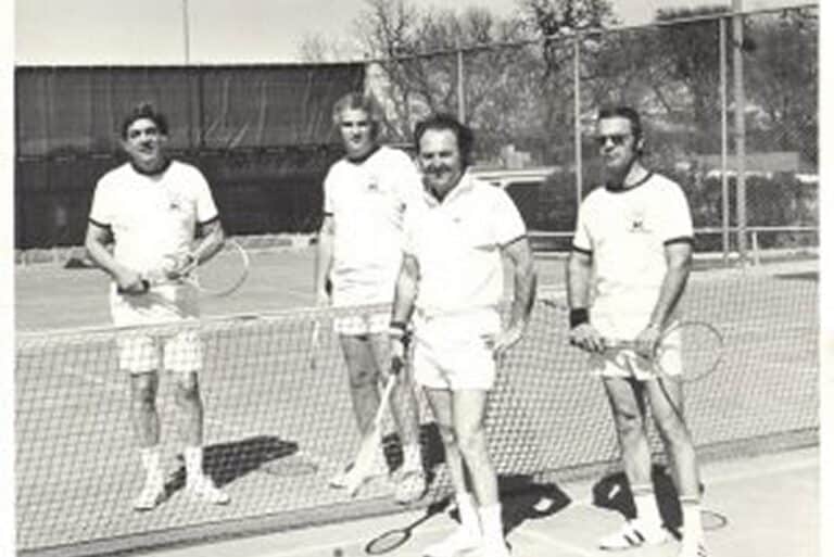 Four men in tennis attire stand on an outdoor court, each holding a racket, posing for a group photo.